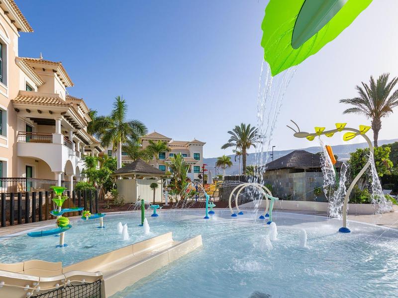 Resortzwembad met watervallen, palmbomen en omliggende gebouwen onder een heldere lucht.