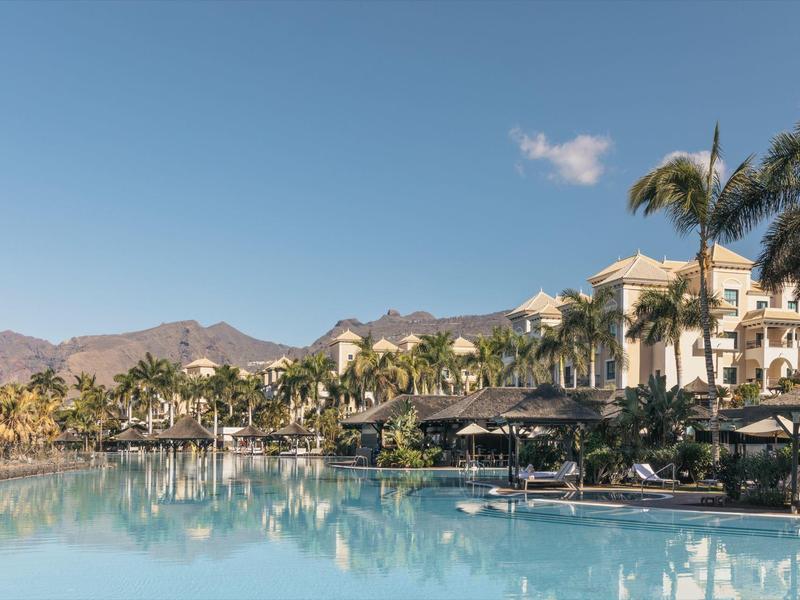 Groot zwembad weerspiegelt palmbomen en resortgebouwen onder een heldere blauwe lucht.