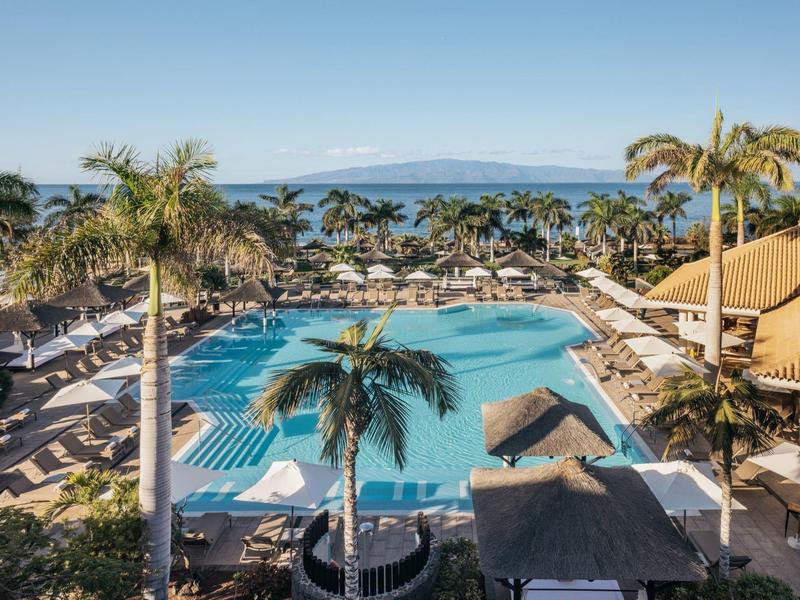 Luxe resort met groot zwembad, ligstoelen, palmbomen en oceaan op de achtergrond onder heldere lucht.