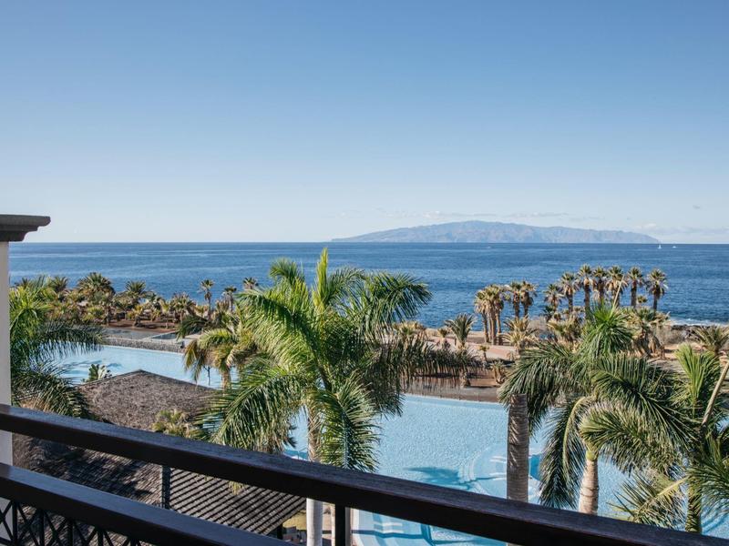 Uitzicht op de oceaan met palmbomen, zwembad en eiland in de verte onder een heldere blauwe lucht.