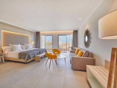 Bright hotel room with large bed, sofa, yellow chairs, and sea view.
