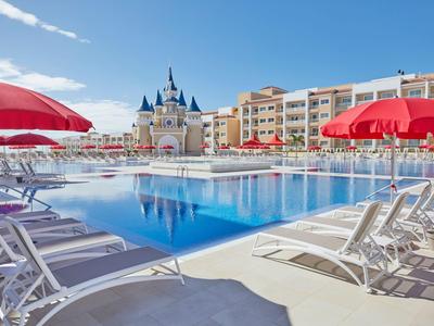 Modern hotel pool with red umbrellas and white lounge chairs under clear sky.