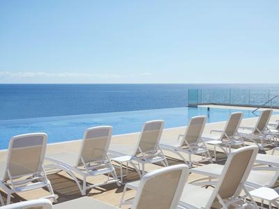 Rows of white lounge chairs by the pool overlooking the sea and clear sky.