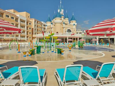 Hotel pool area with blue lounge chairs, water fountains, and rental apartments under blue sky.
