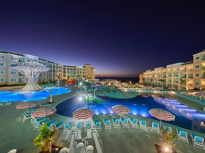 Large hotel resort at night with illuminated pool and sun loungers.