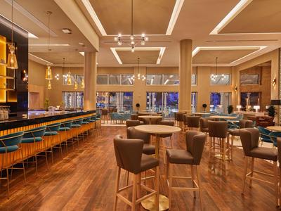 Moderní hotelový bar s dřevěnou podlahou, vysokými stropy, židlemi a stoly.