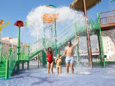 Děti a dospělý si hrají pod vodopádem na vodním hřišti hotelu za slunečného dne.