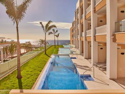 Moderní hotelový komplex s venkovním bazénem, palmami a výhledem na moře při západu slunce.