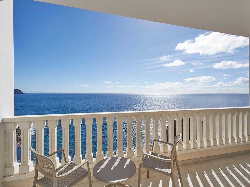 Balkon s bílým zábradlím, židlemi a stolem s výhledem na modré moře pod modrou oblohou.