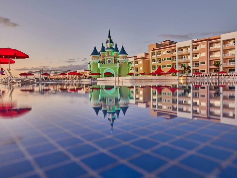 Hotel se zelenou zámkovou architekturou, bazénem a červenými slunečníky při západu slunce.