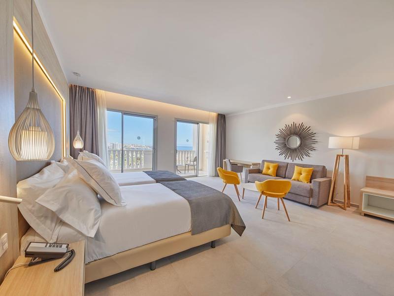 Moderní hotelový pokoj s velkou postelí, posezením se žlutými židlemi a panoramatickým výhledem.
