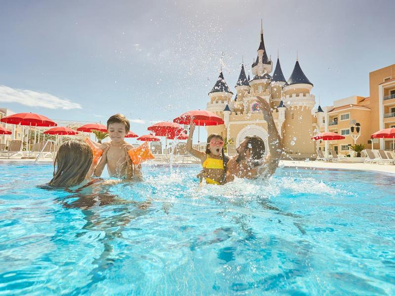 Děti si hrají v bazénu před zámkem s červenými slunečníky a hotelovými budovami v pozadí.