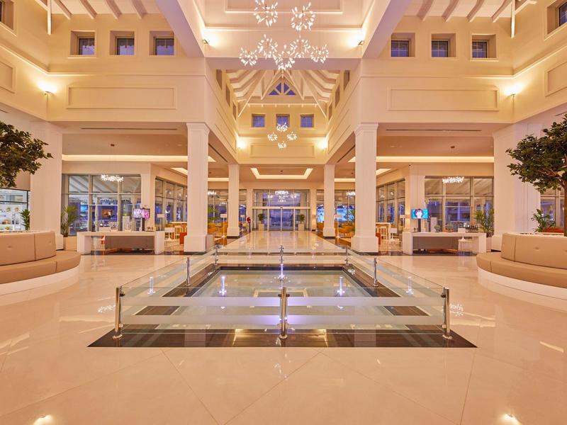 Prostorný světlý hotelový lobby s křišťálovými lustry a stromy v moderních květináčích.