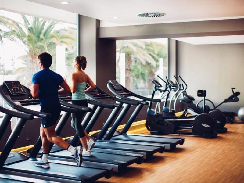 Fitnessstudio mit Laufbändern, zwei Personen joggen, viele Fenster, helle und moderne Einrichtung.