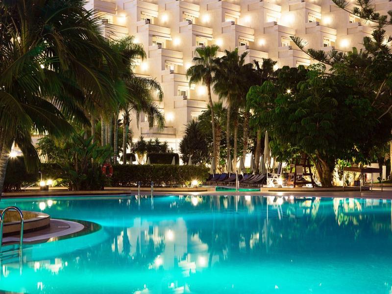 Beleuchteter Resort-Pool bei Nacht mit Palmen und Hotelgebäude im Hintergrund.