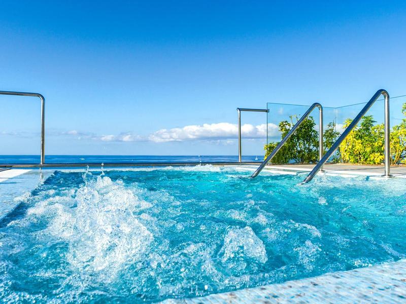 Blauer Whirlpool mit sprudelndem Wasser, Geländer und Meerblick unter klarem Himmel.