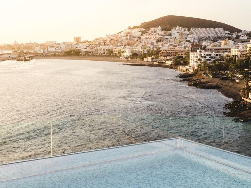 Blick auf Meer mit Küste, weißen Häusern und Hügel im sanften Sonnenlicht, davor ein Pool.