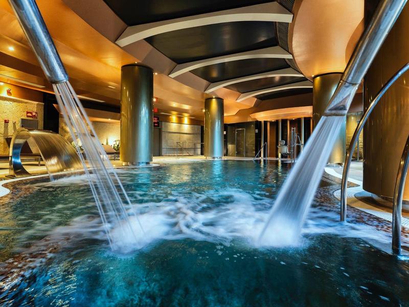 Moderner Hallenbadbereich mit Wasserfontänen, warmem Licht und eleganten Säulen.