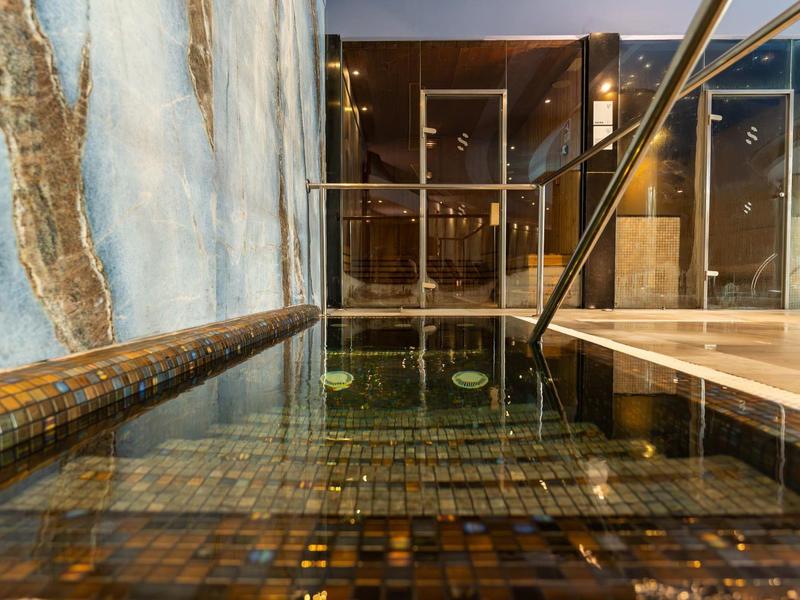 Schwimmbad mit dunklem Wasser, Mosaikfliesen und Glaswand, beleuchtet durch warme Lampen.