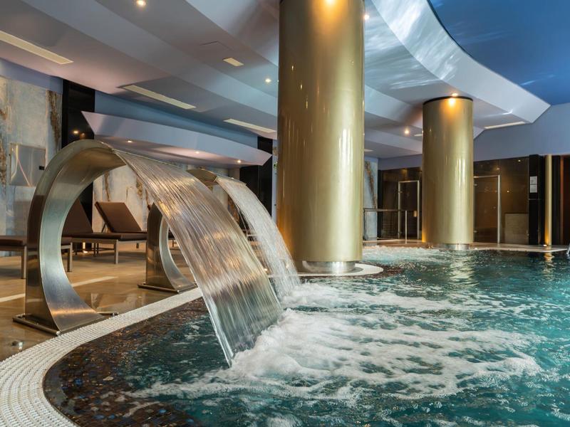 Hallenbad mit Wasserfall-Dusche, goldenen Säulen und sprudelndem Whirlpoolbereich in modernem Design.