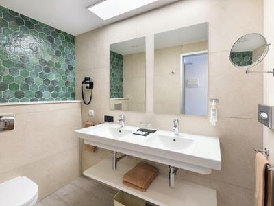 Moderne hotelbadkamer met dubbele wastafels, groene zeshoekige tegels, grote spiegel en beige muren.