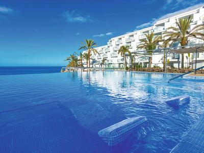 Infinity pool with lounge chairs in front of a modern hotel and blue sky.