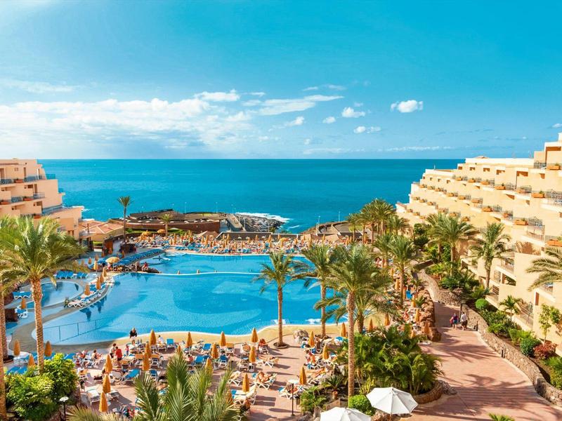 Resort met groot zwembad, palmbomen, ligstoelen en uitzicht op zee onder een deels bewolkte hemel.