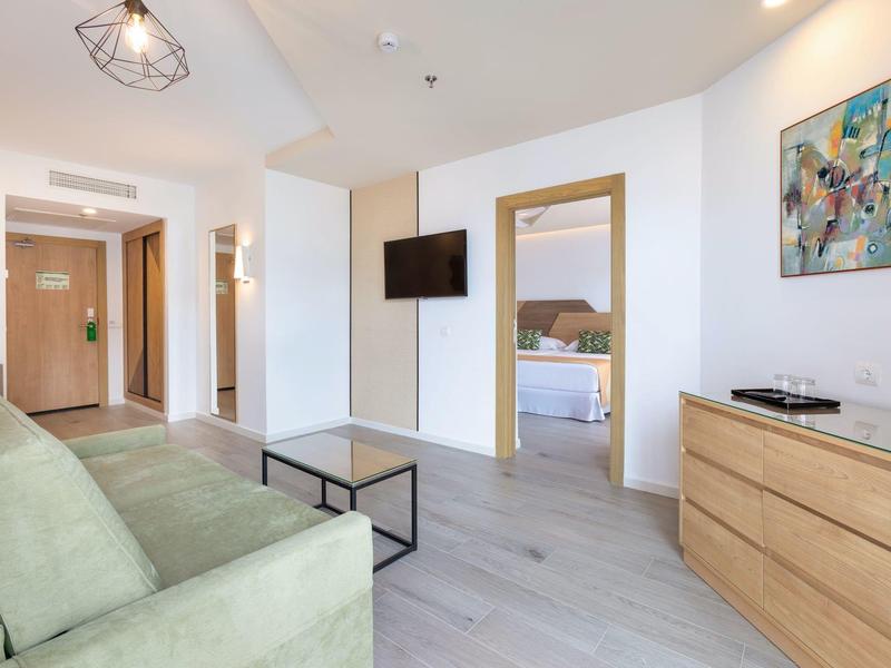 Lichte hotelkamer met groene bank, houten ladekast en tv aan de muur.