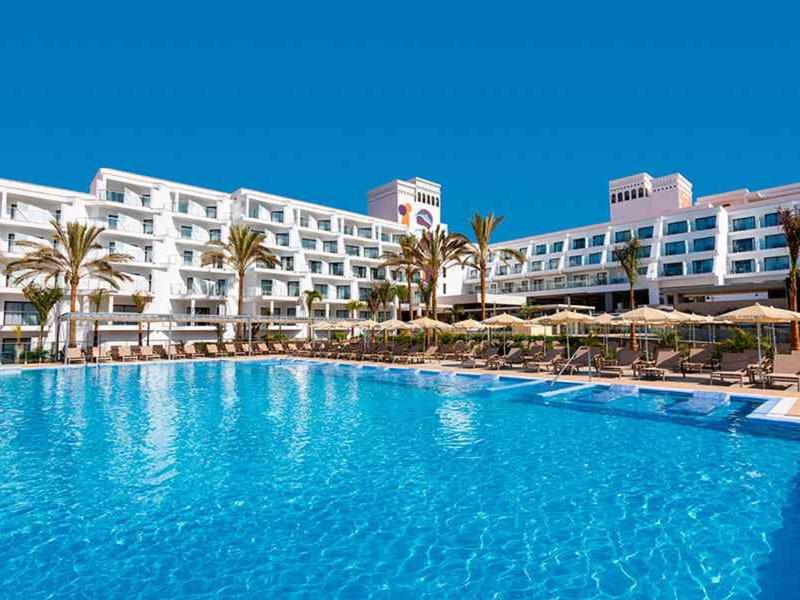 Groot hotel met witte gebouwen rondom een helderblauw zwembad onder een zonnige hemel.