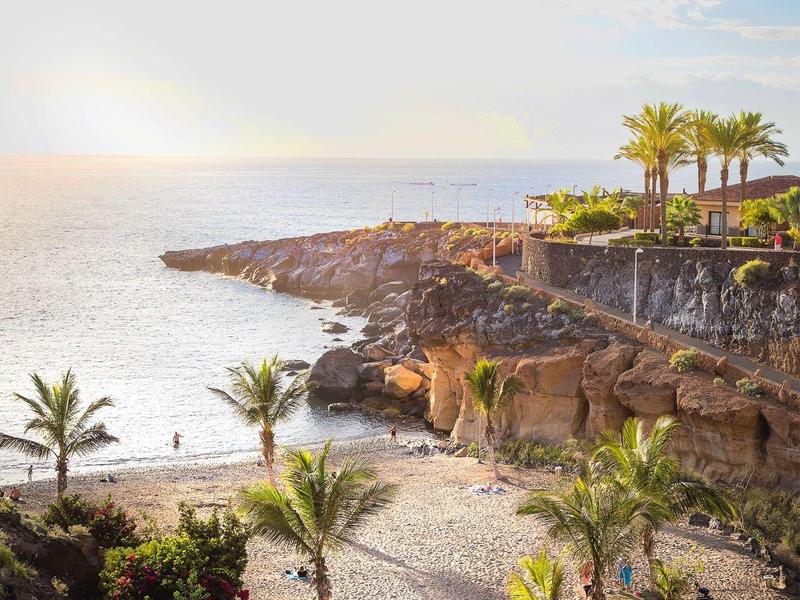 Zonsondergang bij een rotsachtig strand met palmbomen en enkele mensen bij het water.