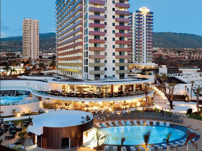 Modern hoogbouwhotel met zwembad en ligstoelen onder een heldere blauwe lucht bij schemering.