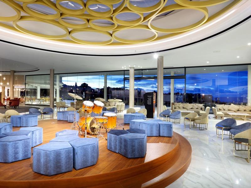 Moderne hotellounge met ronde blauwe ottomans, houten podium en grote glazen ramen met uitzicht op een landschap.