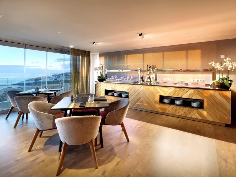 Moderne hotel eetruimte met ronde tafels, beige stoelen en panoramisch uitzicht op de oceaan.