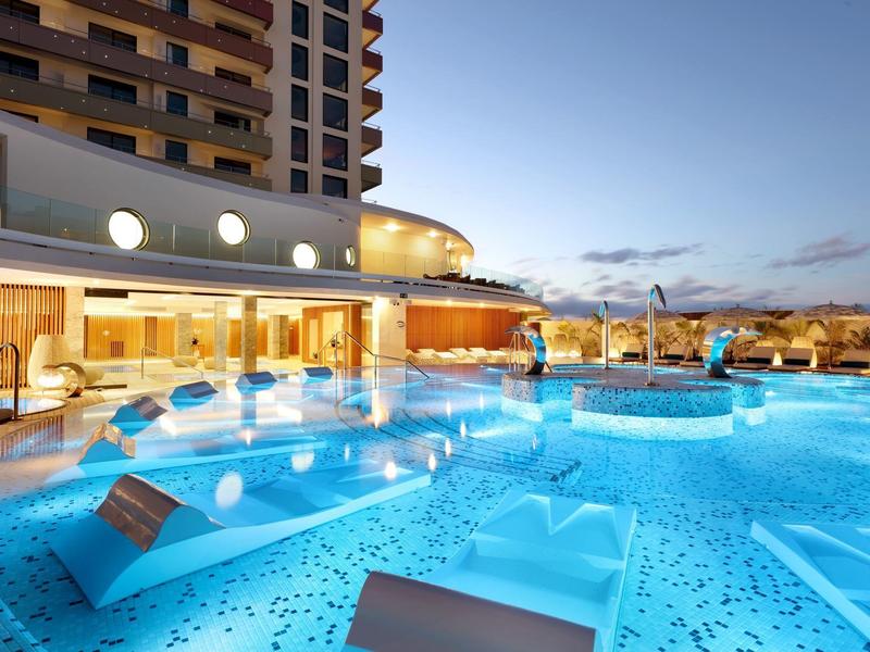 Buitenzwembad van hotel met ligbedden en verlicht water onder een heldere lucht bij zonsondergang.