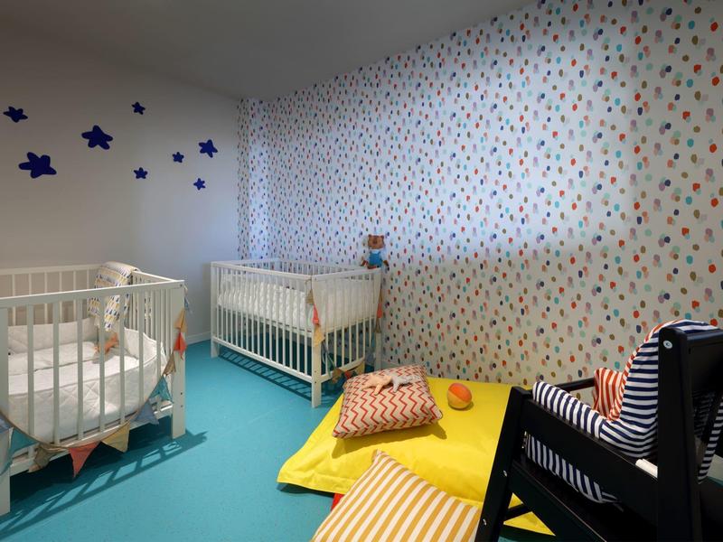 Kleine babykamer met twee witte wiegjes, kleurrijk behang, zachte matten en een zwarte schommelstoel.