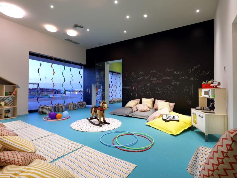 Lichte speelkamer met blauwe vloer, speelgoed, kussens en decoratieve muurkunst.