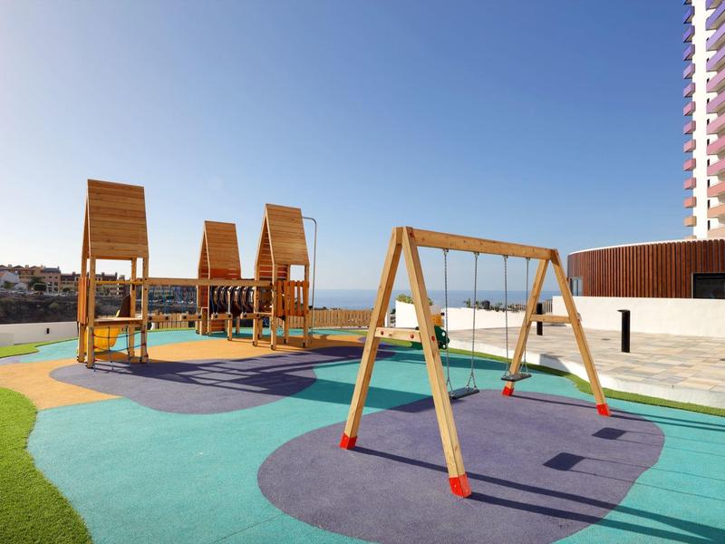 Buitenspeelplaats met houten schommels en klimtoestellen naast een hoog gebouw onder een heldere hemel.