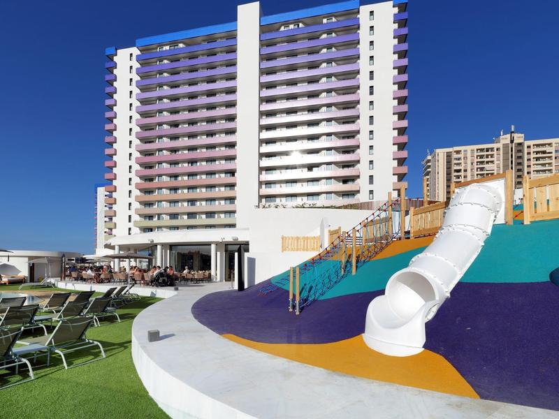 Speelplaats bij hotel met witte buisslide en ligbedden onder een helderblauwe lucht.