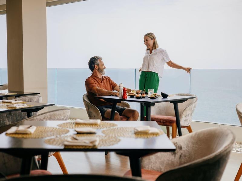 Twee vrouwen genieten van een ontspannen maaltijd op een balkon met uitzicht op zee bij helder weer.