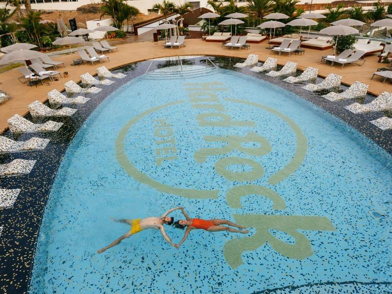 Resortzwembad met ligbedden en parasols omringd door moderne hotelgebouwen onder een bewolkte hemel.