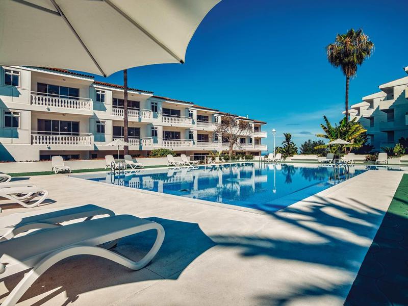 Großer Außenpool mit weißen Liegen, Sonnenschirm und mehrstöckigem Hotel bei klarem Himmel.