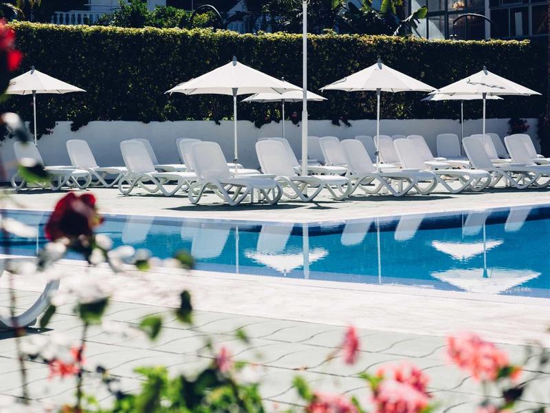 Leerer Poolbereich mit weißen Liegestühlen und Sonnenschirmen im Hintergrund bei Sonnenschein.