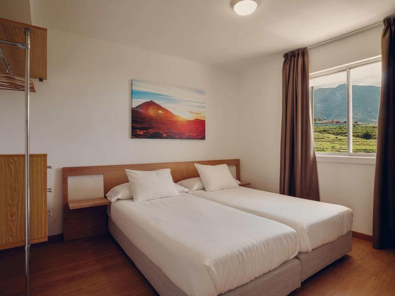 Einfach eingerichtetes Zimmer mit zwei Einzelbetten, hohem Fenster und Landschaftsbild an der Wand.