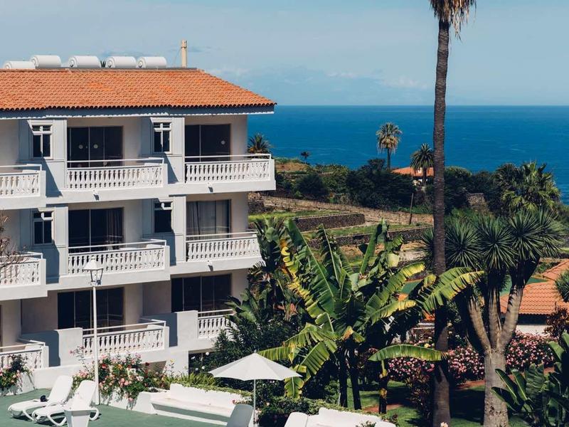 Mehrstöckiges Hotel mit weißem Pool, Liegestühlen und Palmen am Meer unter blauem Himmel.