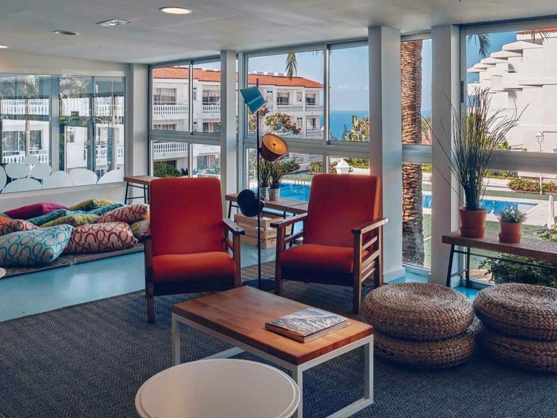 Modernes Wohnzimmer mit großen Fenstern, rotem Sessel, rundem Tisch und bunten Kissen auf Sitzsäcken.