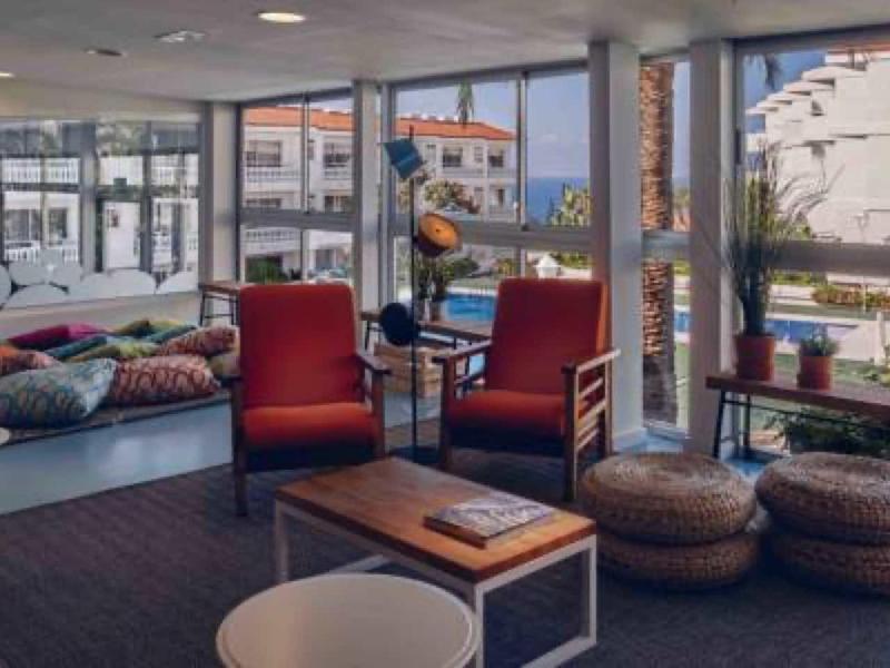 Helles Wohnzimmer mit großen Fenstern, roten Sesseln, bunten Kissen und runden Sitzkissen auf dem Boden.