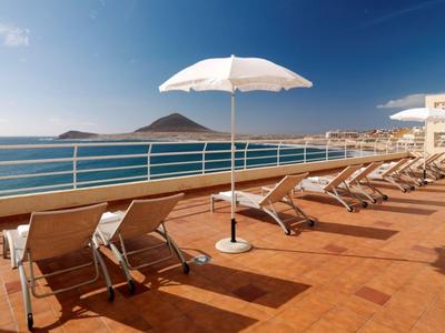 Mehrere Liegestühle und ein Sonnenschirm auf einer Terrasse mit Blick auf das blaue Meer und Inseln.