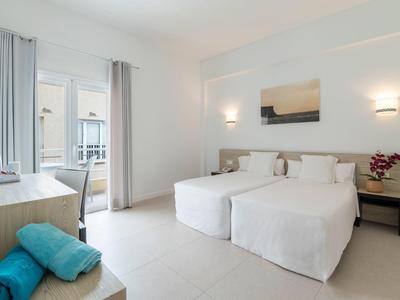 Camera d'hotel luminosa con due letti singoli, scrivania e balcone con luce naturale.