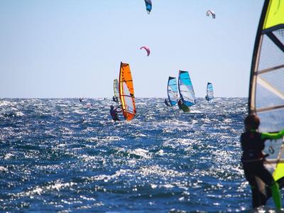 Několik windsurfařů s barevnými plachtami pluje na rozbouřeném moři pod jasnou oblohou.