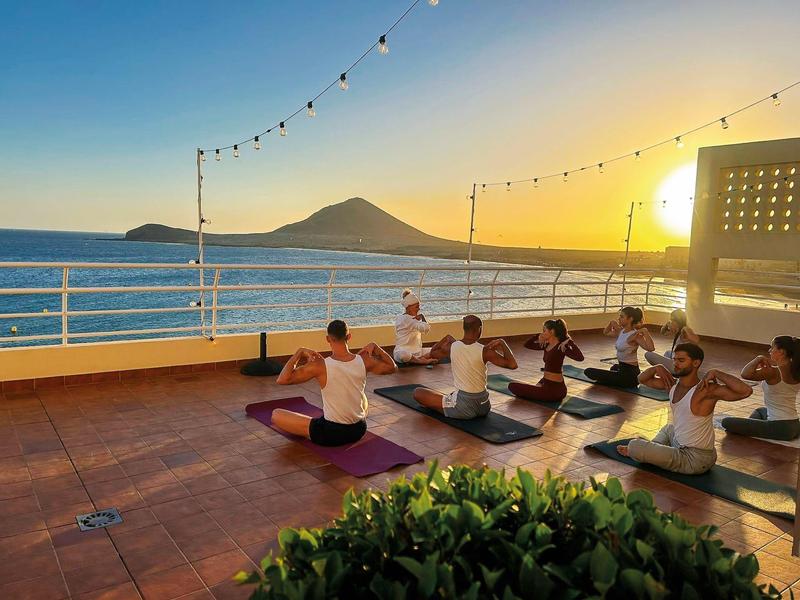 Gruppo che pratica yoga su terrazza panoramica con vista mare e montagna al tramonto.